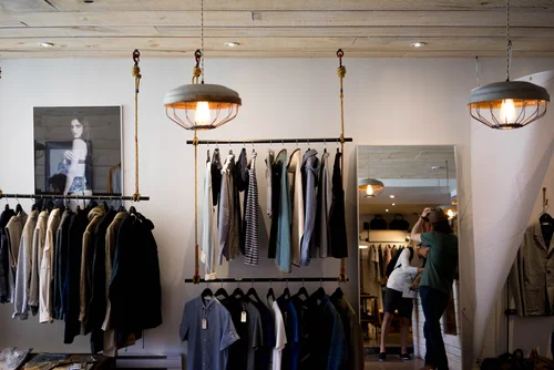 Retail shop interior showcasing clothing racks with various garments, stylish lighting fixtures, and a mirror reflecting shoppers, highlighting commercial renovation and design services by OZ General Contracting.
