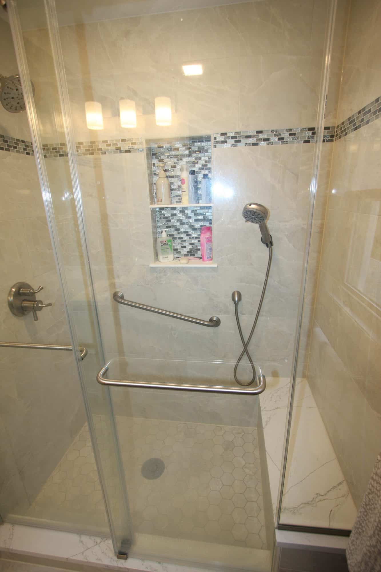 Tall view into the shower showing the mosaic-lined two-shelf niche, handheld shower, grab bars and bench