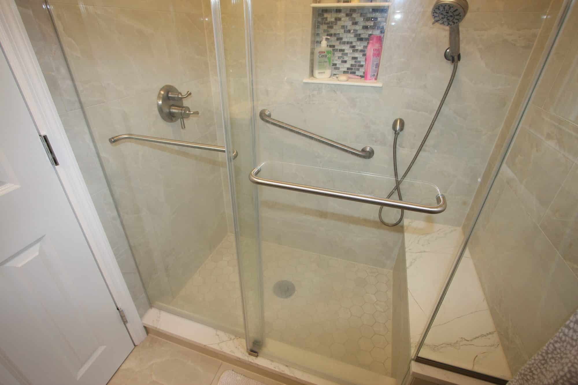 Shower bench with a marble-look top beside a hexagon mosaic shower floor and two brushed nickel grab bars
